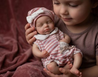 Può includere: Una bambola realistica per bambini vestita con un completo a righe rosa e bianche e un cappello abbinato con un fiocco bianco. La bambola è tenuta da una persona. Gli occhi della bambola sono chiusi e sembra dormire. L'outfit della bambola ha un colletto bianco e finiture in pizzo.