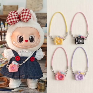 May include: A doll wearing a denim dress and a camera-shaped purse. The purse is pink with a blue bow. The doll has a white bunny hood with a red and white checkered bow. Additional camera-shaped purses in yellow, black, pink, and purple are also shown.
