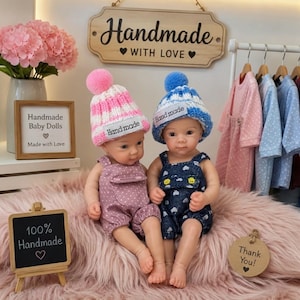 Op de afbeelding: Twee babypoppen met gebreide mutsen met pompons en tuinbroeken. De linkerpop draagt een roze-witte gestreepte muts en een roze tuinbroek met stippen. De rechterpop draagt een blauw-witte gestreepte muts en een donkerblauwe tuinbroek met hartjespatroon. Beide mutsen hebben een wit label met de tekst "Hand made".