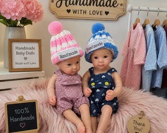 Può includere: Due bambole con cappelli in maglia a pon pon e salopette. La bambola a sinistra indossa un cappello a righe rosa e bianche e una salopette rosa a pois. La bambola a destra indossa un cappello a righe blu e bianche e una salopette blu scuro con motivo a cuori. Entrambi i cappelli hanno un'etichetta bianca con la scritta "Hand made".