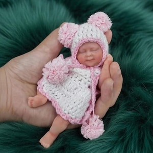 Puede incluir: Una pequeña muñeca bebé vestida con un conjunto de ganchillo blanco y rosa, con un gorro y pompones a juego. La muñeca se sostiene en una mano, destacando su pequeño tamaño y la detallada artesanía de la ropa. La muñeca tiene un tono de piel realista y los ojos cerrados.