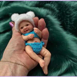 Op de afbeelding: Een miniatuur babypop met een glimlachende uitdrukking, gekleed in een blauwe gehaakte bikini en een witte berenmuts. De pop wordt in een hand gehouden tegen een groene achtergrond.
