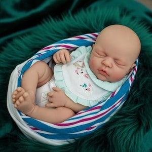 Puede incluir: Una muñeca bebé realista está envuelta en una envoltura a rayas azul, blanca y rosa. La muñeca lleva un babero con un diseño floral y un pañal. El bebé tiene la piel clara y los ojos cerrados, pareciendo dormido. El fondo es una superficie de felpa verde oscuro.
