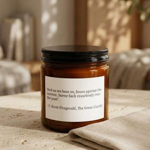May include: A brown glass jar with a black lid, containing a candle. A white label on the jar features a quote from F. Scott Fitzgerald's "The Great Gatsby". The candle sits on a light-colored surface with a folded cloth.