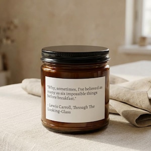 May include: A brown glass jar candle with a black lid. A white label on the jar features a quote from Lewis Carroll's "Through The Looking-Glass". The quote reads: "Why, sometimes, I've believed as many as six impossible things before breakfast."