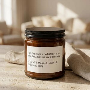 Peut inclure: Une bougie en pot de verre marron avec un couvercle noir. La bougie porte une étiquette blanche avec la citation "To the stars who listen - and the dreams that are answered" et le nom de l'auteur. La bougie est sur une surface claire.