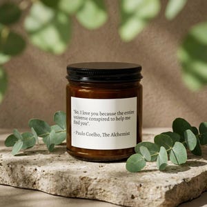 Peut inclure: Une bougie en pot de verre brun avec un couvercle noir et une étiquette blanche avec une citation de Paulo Coelho. La bougie est entourée de feuilles d'eucalyptus vertes sur une surface en pierre.