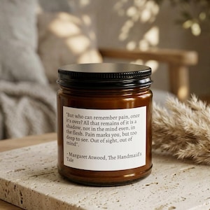 Peut inclure: Une bougie en pot de verre brun avec un couvercle noir. La bougie porte une étiquette blanche avec un texte de "The Handmaid's Tale" de Margaret Atwood. La bougie repose sur une surface en pierre de couleur claire.