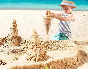 May include: A child wearing a straw hat and striped tank top builds a sandcastle on a sandy beach. The child is using a cone-shaped tool to pour sand, creating a castle with two towers and a surrounding wall. The ocean is visible in the background.