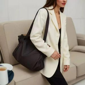 May include: A dark brown tote bag with a shoulder strap. The bag is made of a soft material and has a gathered design. The person is wearing a white blazer and black clothing.