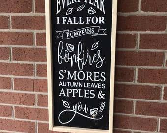 May include: A rectangular wooden sign with a black background and a light-colored wood frame. The sign features white text that reads: EVERY YEAR I FALL FOR PUMPKINS, bonfires, S'MORES, AUTUMN LEAVES, APPLES & you, decorated with small leaf icons.