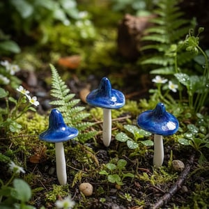Puede incluir: Tres setas de cerámica con sombreros azul oscuro brillante y tallos blancos se encuentran en un lecho de musgo verde y follaje. Las setas en miniatura son una decoración de jardín caprichosa, que añade un toque de fantasía.