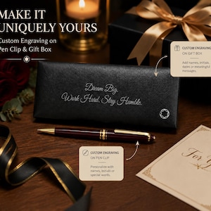 May include: A burgundy ballpoint pen with gold-tone accents and a clip, positioned in front of a black rectangular gift box featuring the engraved text "Dream Big. Work Hard. Stay Humble." and a small circular logo on the bottom right corner.