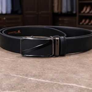 May include: A black belt featuring a smooth, matte finish strap and a rectangular, dark metallic buckle with a polished, reflective center panel. The belt is coiled, showcasing a clean, solid design with a matching black leather loop.