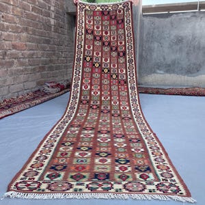 Könnte beinhalten: Ein langer, rechteckiger Läufer mit einem sich wiederholenden Gittermuster aus geometrischen Motiven in Rot-, Blau-, Grün- und Cremetönen. Der Teppich hat eine dekorative Bordüre mit kreisförmigen Mustern und weiße Fransen an den Enden.