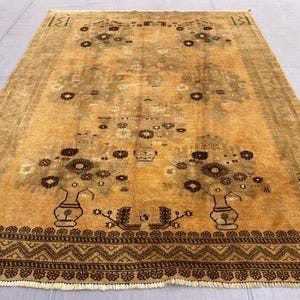 May include: A rectangular, golden-yellow rug with a floral design. The rug features two vases with flower arrangements, surrounded by smaller floral motifs. The border is a dark brown with a geometric pattern. The rug is likely made of wool or a similar material.