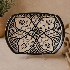 Peut inclure: Un plateau ovale en céramique présentant une base de couleur crème avec des motifs noirs complexes peints à la main. Le design comprend un motif floral central, des formes géométriques en losange et des bordures pointillées, le tout encadré par un bord noir.