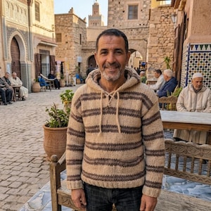 Peut inclure: Un pull à capuche à manches longues avec des rayures horizontales alternant entre le beige et le brun foncé. Le vêtement présente une texture en maille épaisse, une capuche à cordon de serrage et une poche kangourou, porté sur un jean bleu foncé.