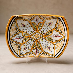 Peut inclure: Un plat de service rectangulaire en céramique aux bords arrondis, présentant un motif floral central jaune et blanc avec des accents orange, rouges et verts, entouré d'une bordure jaune à texture pointillée et contours noirs.