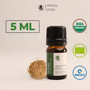 May include: A 5 ml amber glass bottle of vetiver essential oil with a black cap and a white label featuring the text Vetiver, 100% Tinh Dầu Hữu Cơ, and 0.17 fl oz. A small bundle of dried vetiver roots sits beside the bottle, with USDA Organic icons.