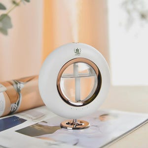 May include: A circular white humidifier with a rose gold inner ring and a clear central water reservoir. The device features a small green logo at the top and a vertical white misting tube inside. It stands on a small, round rose gold base.