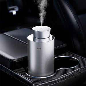 Può includere: Un diffusore per auto cilindrico color argento con finitura metallica, dotato di un ugello superiore che emette un flusso costante di nebbia bianca. Il dispositivo presenta una piccola spia orizzontale sulla parte anteriore ed è in un portabicchieri.