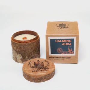 May include: A cylindrical candle in a bark-like container with a wooden wick, next to a box with the text "CALMING AURA". The candle's lid is also visible, featuring a logo and the brand name "Le Restique".