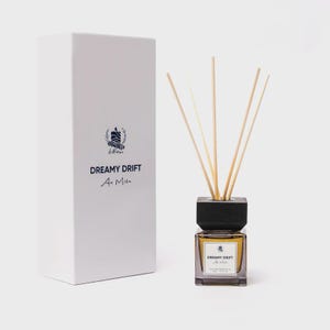 May include: A reed diffuser set featuring a square glass bottle with a dark wooden cap and five light-colored reeds. The bottle has a white label reading DREAMY DRIFT An Miên. Beside it stands a tall white rectangular box with the same branding.