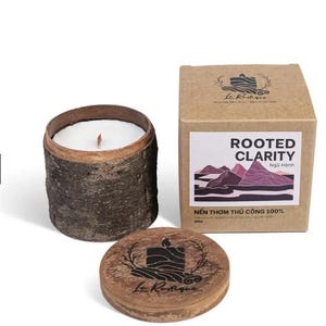 Puede incluir: Una vela blanca en un recipiente cilíndrico de madera con textura de corteza natural, acompañada de una tapa de madera a juego y una caja de cartón cuadrada. La etiqueta muestra el texto ROOTED CLARITY, un gráfico de montaña y 100g.