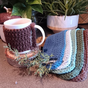 Può includere: Una tazza di ceramica bianca con una custodia all'uncinetto marrone e un involucro per manico abbinato poggia su un sottobicchiere di legno. Una pila di sottobicchieri all'uncinetto nei toni del blu, verde e marrone è accanto alla tazza. Il verde aggiunge un tocco naturale.