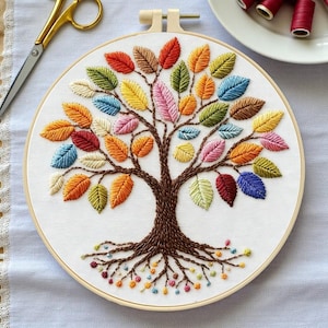 May include: Embroidered tree art on white fabric, featuring a brown trunk and roots, with colorful leaves in shades of red, orange, yellow, green, blue, and pink. The embroidery is set in a wooden hoop, with gold scissors and spools of thread in the background.