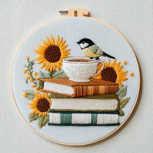 Op de afbeelding: Geborduurde hoepelkunst met een stapel boeken, zonnebloemen en een theekopje met een vogel erbovenop. De boeken zijn bruin, groen en wit. De zonnebloemen zijn geel en bruin. De vogel is zwart, wit en geel. De achtergrond is lichtblauw.