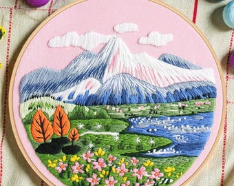 Op de afbeelding: Geborduurde landschapsscène in een houten ring. Het ontwerp toont besneeuwde bergen, een blauwe rivier, groene velden, oranje bomen en roze en gele bloemen, geplaatst op een roze stoffen achtergrond.