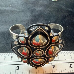 Pu&ograve; includere: Un bracciale a fascia con un'ampia banda in metallo argentato martellato e un grande elemento centrale nero decorato. Il centro &egrave; ornato da molteplici cabochon argentati riflettenti di forma irregolare e piccoli strass trasparenti.