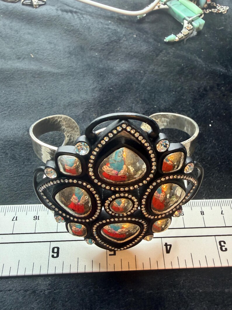 Pu&ograve; includere: Un bracciale a fascia con un'ampia banda in metallo argentato martellato e un grande elemento centrale nero decorato. Il centro &egrave; ornato da molteplici cabochon argentati riflettenti di forma irregolare e piccoli strass trasparenti.