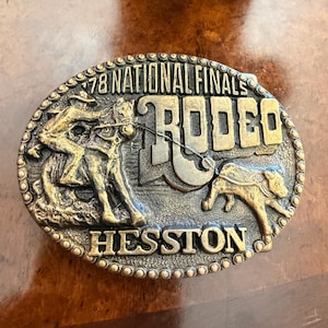 Puede incluir: Una hebilla de cinturón ovalada de tono latón con un diseño en relieve de una persona montando un caballo y un ternero. El texto dice 78 NATIONAL FINALS RODEO y HESSTON. El borde está decorado con una serie de pequeñas cuentas circulares.
