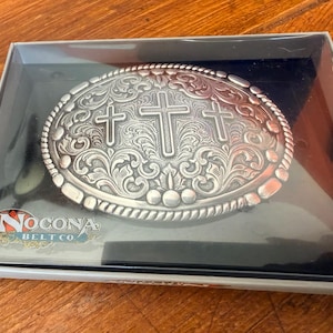 May include: An oval silver-toned belt buckle featuring an embossed floral scroll pattern and three central crosses. The buckle is housed in a black display box with the Nocona Belt Co. logo printed in the lower left corner of the clear plastic lid.