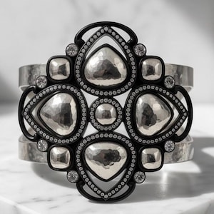 Pu&ograve; includere: Un bracciale a fascia larga con una disposizione centrale di forme a goccia e circolari in argento martellato incastonate in una cornice nera. La cornice nera &egrave; accentuata da piccole pietre chiare e il bracciale ha una finitura argento spazzolato.