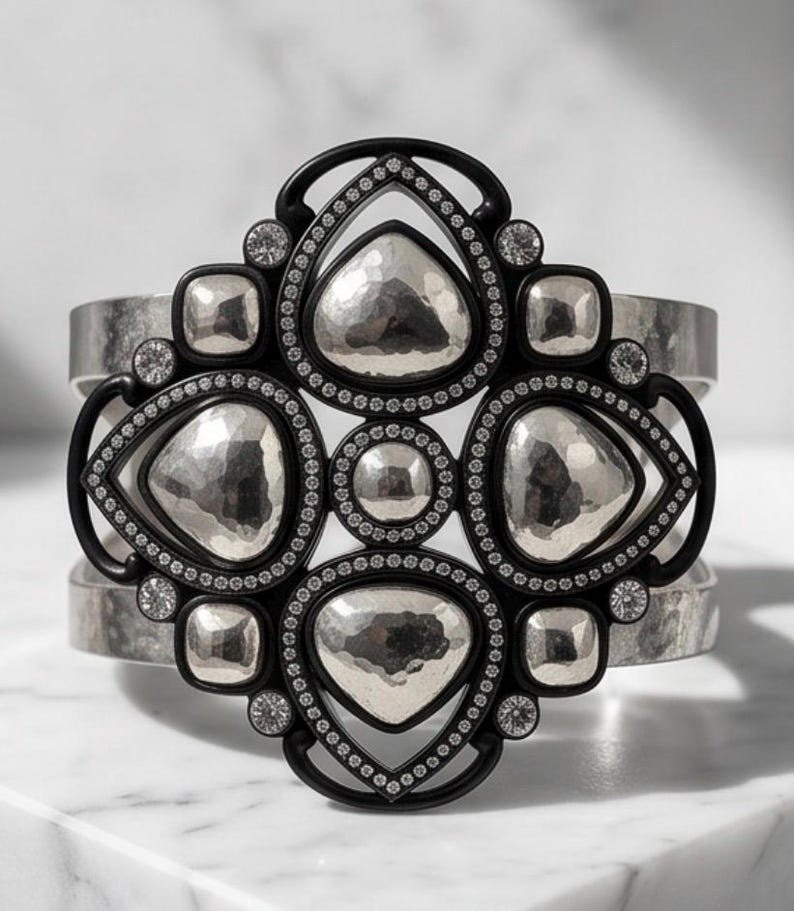 Pu&ograve; includere: Un bracciale a fascia larga con una disposizione centrale di forme a goccia e circolari in argento martellato incastonate in una cornice nera. La cornice nera &egrave; accentuata da piccole pietre chiare e il bracciale ha una finitura argento spazzolato.