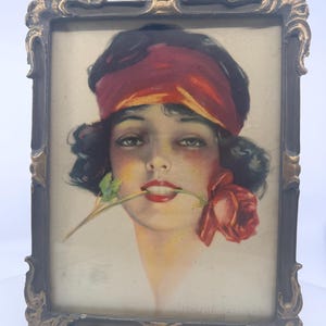May include: Vintage portrait of a woman with dark hair and a red headband, holding a red rose in her mouth. The artwork is framed in a dark, ornate frame with gold accents. The image has a soft, vintage aesthetic.