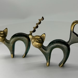 Puede incluir: Dos herramientas de metal con forma de gato, acabado en tono latón y pátina oscura. Un gato tiene una cola en espiral de sacacorchos, mientras que el otro tiene una cola en bucle triangular que funciona como abridor de botellas. Lomo arqueado.