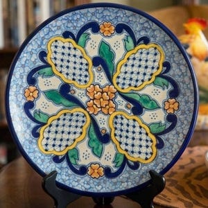 May include: A round ceramic plate featuring a central cluster of orange flowers surrounded by four yellow-bordered, blue-and-white grid patterned shapes. The design includes green leaves and dark blue scrollwork on a light blue speckled background.