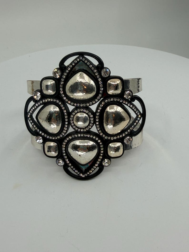 Pu&ograve; includere: Un bracciale a fascia larga caratterizzato da una cornice nera centrale con una disposizione geometrica di placche in metallo argentato martellato. La cornice nera &egrave; impreziosita da piccoli strass trasparenti e il bracciale &egrave; in metallo argentato lucido.
