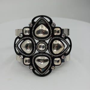 Pu&ograve; includere: Un bracciale a fascia larga caratterizzato da una cornice centrale nera con una disposizione geometrica di inserti in metallo argentato martellato. Il design &egrave; impreziosito da piccoli strass trasparenti lungo i bordi neri a contrasto.