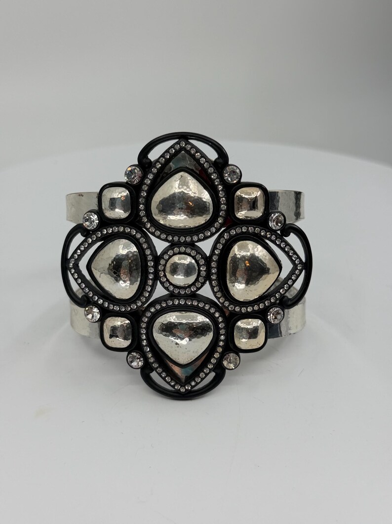 Pu&ograve; includere: Un bracciale a fascia larga caratterizzato da una cornice centrale nera con una disposizione geometrica di inserti in metallo argentato martellato. Il design &egrave; impreziosito da piccoli strass trasparenti lungo i bordi neri a contrasto.