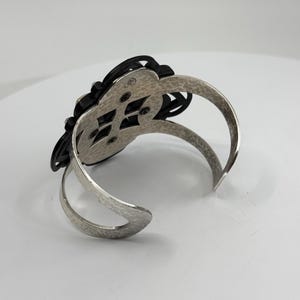 Pu&ograve; includere: Un bracciale rigido color argento con una finitura in metallo martellato e testurizzato sulla fascia. Il pezzo centrale &egrave; un ornamento nero traforato a forma di fiore, fissato al telaio in metallo con quattro viti visibili.