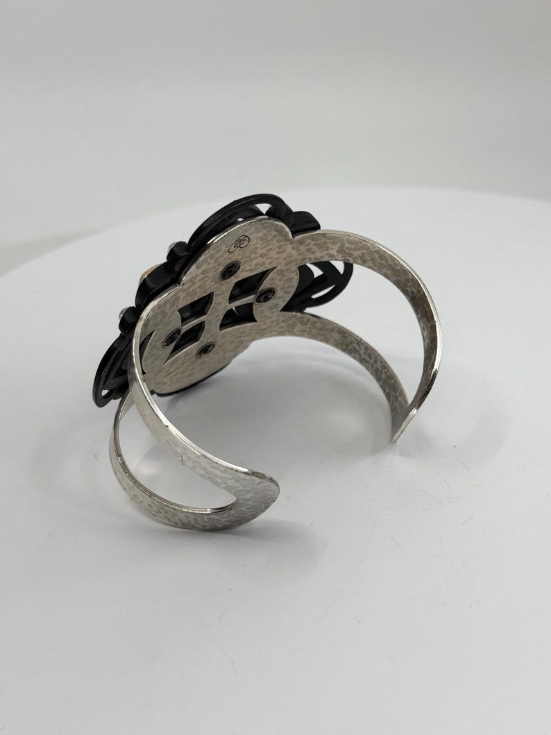 Pu&ograve; includere: Un bracciale rigido color argento con una finitura in metallo martellato e testurizzato sulla fascia. Il pezzo centrale &egrave; un ornamento nero traforato a forma di fiore, fissato al telaio in metallo con quattro viti visibili.