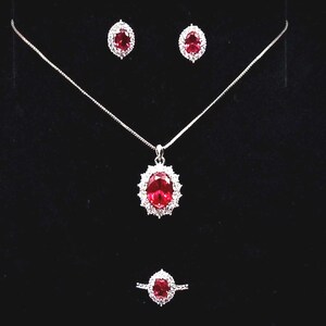 May include: A jewelry set consisting of a necklace with an oval red gemstone pendant surrounded by small clear stones, matching oval red gemstone stud earrings, and a matching oval red gemstone ring, all set in a silver-toned metal frame.