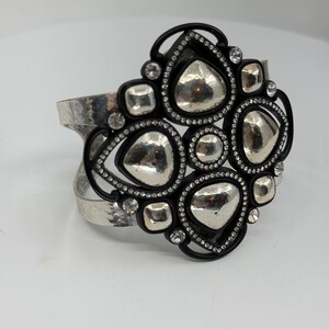 Pu&ograve; includere: Un bracciale a fascia larga con una banda in metallo argentato martellato. Il pezzo centrale &egrave; un'ornata cornice nera contenente molteplici forme argentate lisce e irregolari e piccoli cristalli trasparenti disposti in un motivo.