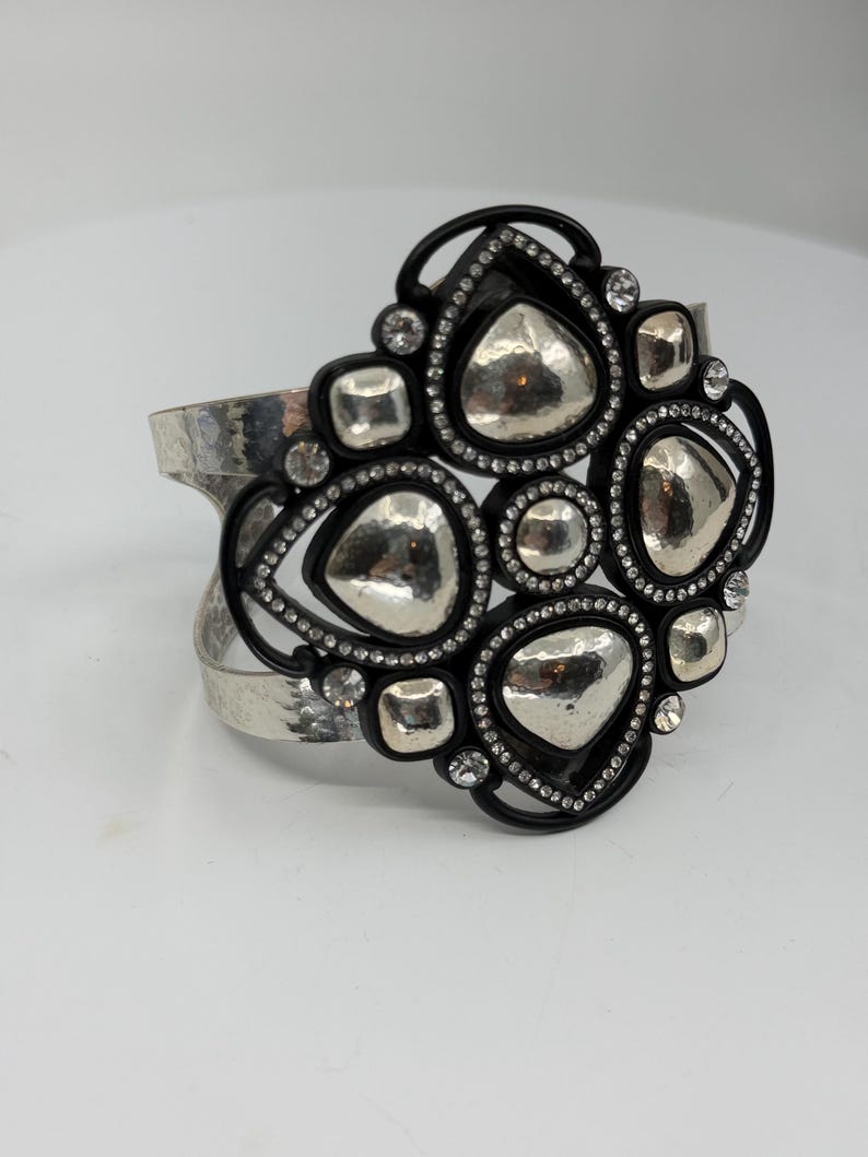 Pu&ograve; includere: Un bracciale a fascia larga con una banda in metallo argentato martellato. Il pezzo centrale &egrave; un'ornata cornice nera contenente molteplici forme argentate lisce e irregolari e piccoli cristalli trasparenti disposti in un motivo.